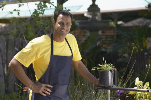 A man wearing a bright yellow t-shirt and a dark blue apron is standing in a well-maintained garden, smiling at the camera while holding a small black plant pot containing a young, green plant. The garden features a lush, green lawn with neatly trimmed grass in the foreground, bordered by a variety of shrubs, flowering plants, and ornamental grasses. Behind him, there is a wooden fence and a garden shed partially obscured by foliage. In the background, mature trees and a multi-tiered stone water fountain are visible, adding a focal point to the outdoor space. The area appears to be well-lit by natural daylight, with some shade provided by a canopy or pergola overhead. The setting suggests routine gardening and outdoor maintenance work, consistent with professional garden services in Purley, and the scene reflects a healthy, landscaped backyard suitable for outdoor activities and relaxation.