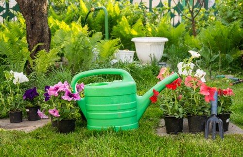 A well-maintained garden scene showing a small area of lush, green grass bordered by a variety of potted flowering plants in shades of purple, pink, and white, including petunias and begonias. In the background, there is a mix of mature trees and dense greenery, providing natural shade and privacy. A white ceramic or stone planter is partially visible among the shrubs, and a garden hose with a red nozzle is resting on the soil near a gardening fork. The garden features a natural, informal layout with a balance of flowering plants and shaded foliage, suggesting regular outdoor maintenance typical of professional gardening services in Purley. The overall atmosphere appears bright and inviting, indicating a clear day with natural sunlight enhancing the vibrant colours of the plants and the lushness of the lawn, suitable for outdoor leisure. This scene reflects a typical cared-for garden that might benefit from routine lawn care, planting, and maintenance services offered by Gardening Services Purley.