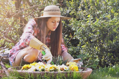 A young woman with long brown hair, wearing a straw hat, a red and white checkered shirt, and gardening gloves, is kneeling in a lush garden. She is carefully tending to a cluster of flowering plants with yellow and white blooms, positioned in a circular bed bordered by soil and green grass. Behind her, dense foliage and trees provide a shaded backdrop, indicating a well-maintained outdoor space likely part of a residential garden in Purley. The scene is illuminated by natural daylight, suggesting a pleasant, sunny day suitable for gardening activities. This outdoor environment features a mix of vibrant flowers, healthy grass, and shrubbery, highlighting professional gardening and lawn care potential services offered by Gardening Services Purley in the local area, including plant tending, soil maintenance, and garden landscaping efforts.