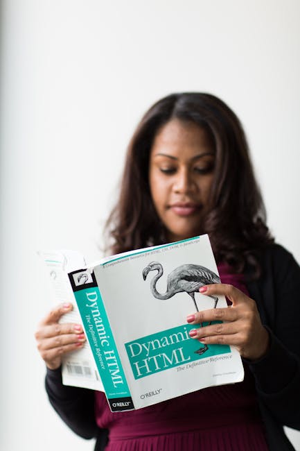 The image shows a woman with dark, wavy hair wearing a black blazer over a burgundy top, holding a book titled 'Dynamic HTML' with a flamingo illustration on the cover. She appears to be reading or ex