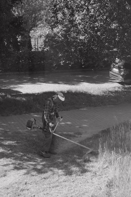 A person wearing a cap and checkered shirt engaging in garden maintenance by using a rake or similar tool to clear overgrown grass and weeds along a garden border. The garden features a lawn area with